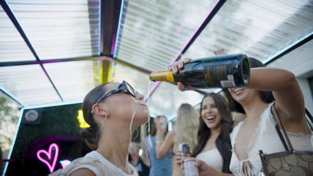 Group of friends at an outdoor neon-lit canopy party; one friend leans back as another pours champagne into her mouth while others laugh.