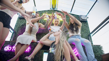 Group of young women on a rooftop patio toasting and drinking canned cocktails under a neon sign