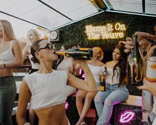Sunset rooftop party with women celebrating; woman in crop top and sunglasses drinks champagne from a bottle under a glowing neon sign.
