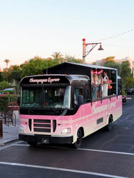 Pink 'Champagne Express' party bus with open rooftop bar and people on deck, parked in a palm-lined plaza at sunset