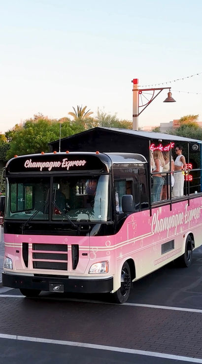 Pink 'Champagne Express' party bus with open rooftop bar and people on deck, parked in a palm-lined plaza at sunset