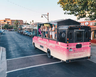Pink open-air party bus with guests on an upper deck driving through a downtown street at sunset, framed by string lights, palm trees, shops, and parked cars.