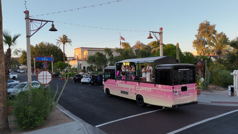 Pink open-air party bus with people onboard rolling through a palm‑lined downtown street at sunset, string lights overhead, cacti and parked cars along the sidewalk.
