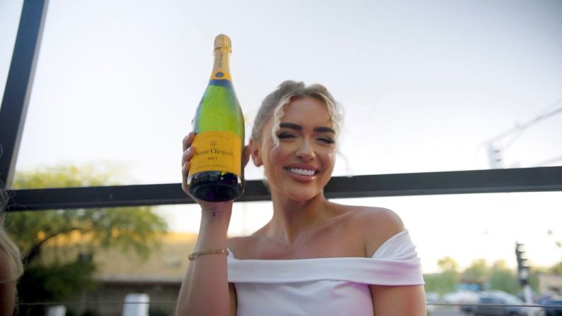 Smiling woman in an off-shoulder white top holding a large champagne bottle aloft on an outdoor patio at dusk with a blurred city street and trees in the background.