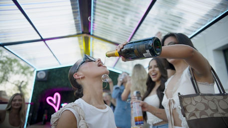 Group of women at an urban rooftop patio party, one pouring champagne into another’s mouth under a transparent canopy with neon heart lighting