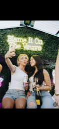 Two women taking a selfie at a lively event photo wall — neon “Blame It On The Veuve” sign on a green hedge backdrop, holding cans and a bottle of sparkling wine, wearing casual summer outfits.
