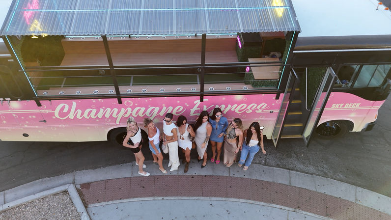 Aerial view of a group of women in party outfits posing beside a pink champagne-themed party bus parked at a curb, giving a festive night-out vibe.
