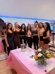 Seven women in workout wear posing at a barre-style fitness studio for a bachelorette party — bride-to-be with pink sash and heart sunglasses, champagne and glasses, and a floral centerpiece on a pink-covered table.