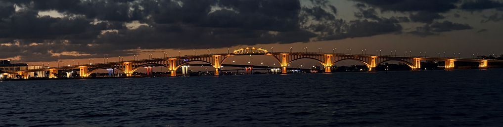 Golden-lit arched bridge glowing at dusk over dark harbor waters, dramatic clouds overhead and city lights reflecting on the rippled surface.