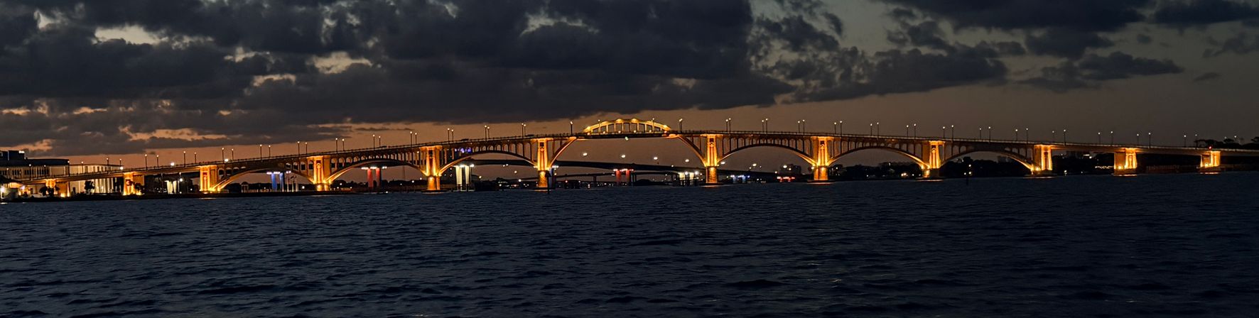 Golden-lit arched bridge glowing at dusk over dark harbor waters, dramatic clouds overhead and city lights reflecting on the rippled surface.