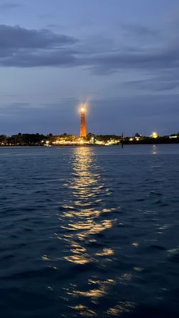 Golden-beacon lighthouse at dusk, its light slicing across calm harbor waters and reflecting in rippled waves beneath a moody twilight sky.