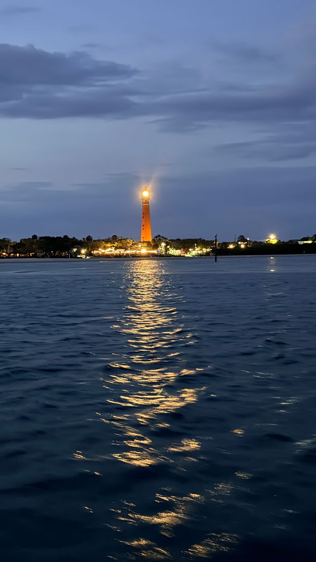Golden-beacon lighthouse at dusk, its light slicing across calm harbor waters and reflecting in rippled waves beneath a moody twilight sky.