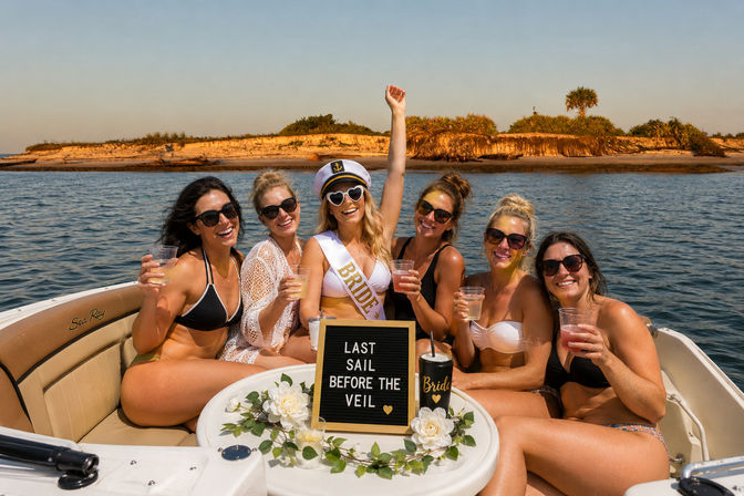 Bachelorette boat party: bride-to-be in captain hat and sash with friends on a coastal boat, drinks raised, ’Last sail before the veil’ sign and sandy island shoreline