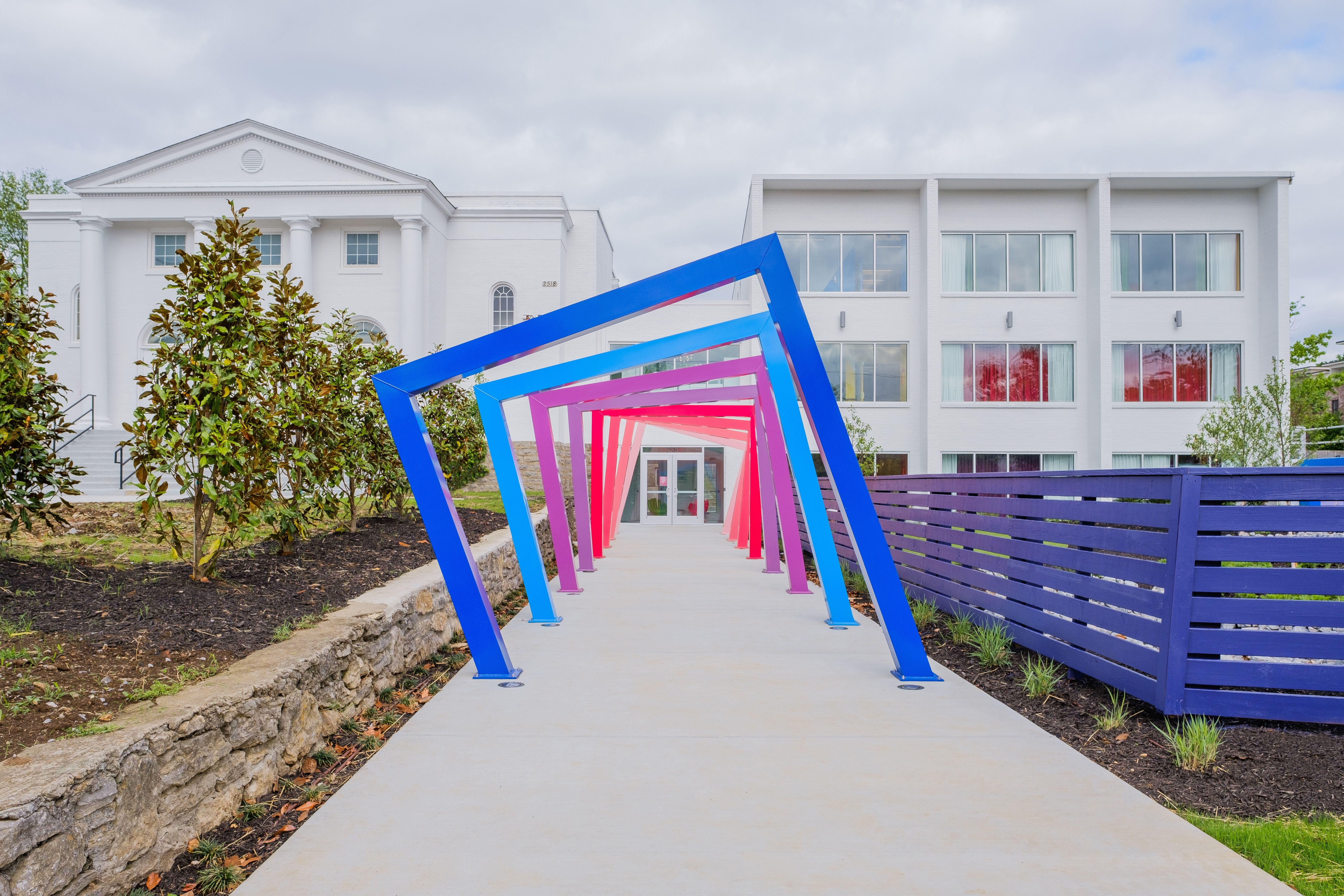 Vibrant angled metal frame tunnel in blue, purple and pink over a concrete path leading to a white modern building entrance, flanked by a purple slatted fence and landscaped planting beds
