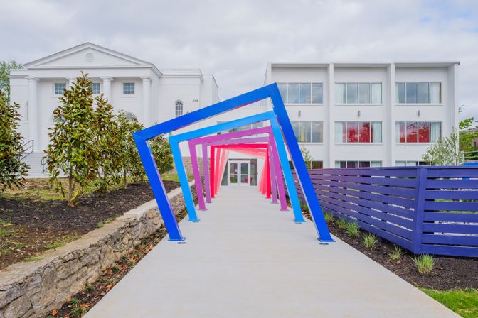 Vibrant angled metal frame tunnel in blue, purple and pink over a concrete path leading to a white modern building entrance, flanked by a purple slatted fence and landscaped planting beds