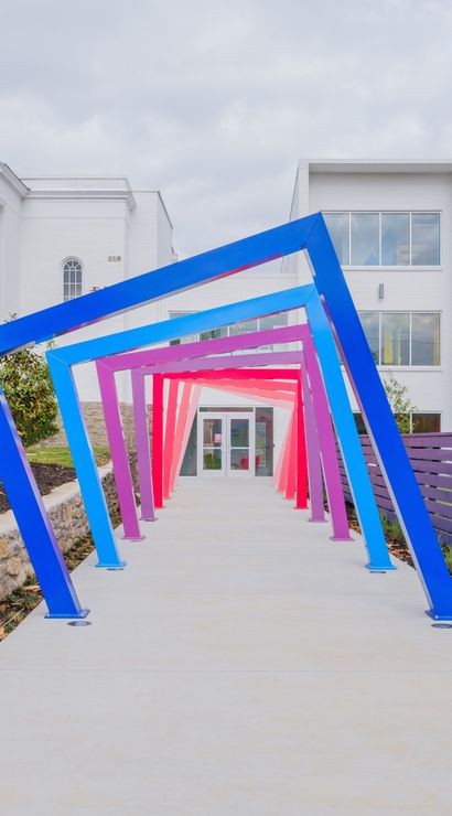 Vibrant angled metal frame tunnel in blue, purple and pink over a concrete path leading to a white modern building entrance, flanked by a purple slatted fence and landscaped planting beds