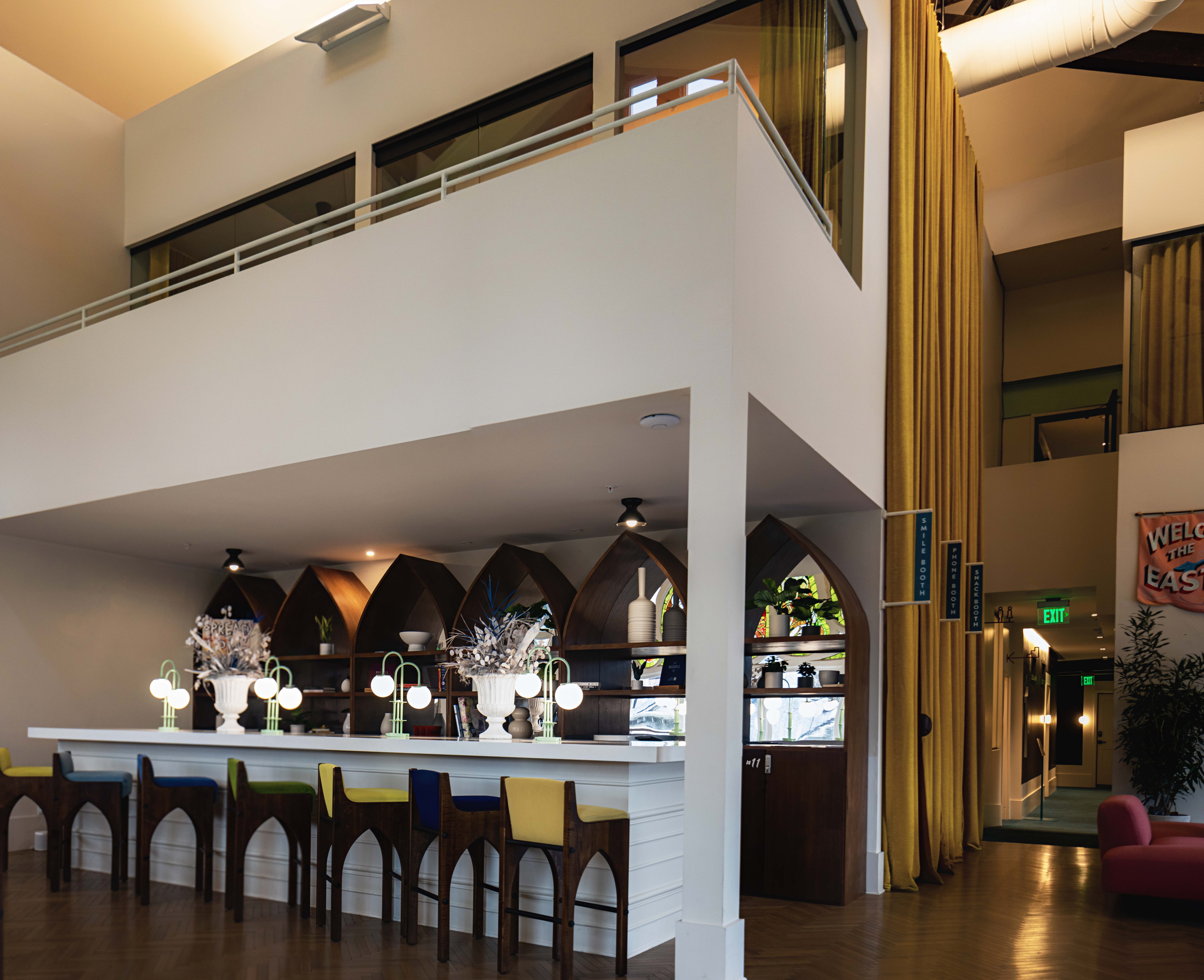 Bright modern hotel lobby bar interior with high mezzanine, arched wooden shelves, colorful bar stools, glowing globe lamps and tall yellow curtains