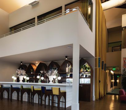 Bright modern hotel lobby bar interior with high mezzanine, arched wooden shelves, colorful bar stools, glowing globe lamps and tall yellow curtains