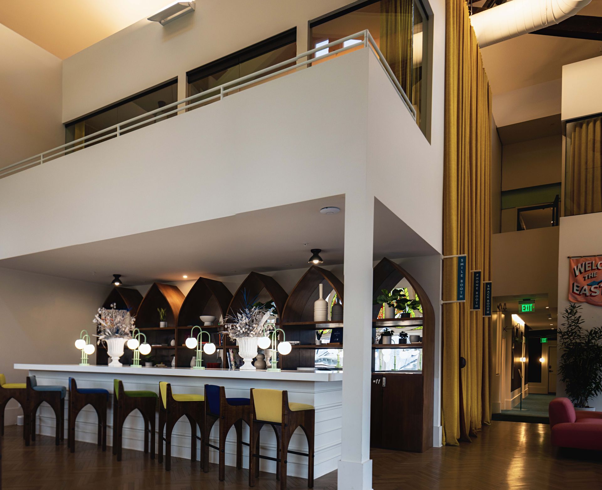 Bright modern hotel lobby bar interior with high mezzanine, arched wooden shelves, colorful bar stools, glowing globe lamps and tall yellow curtains