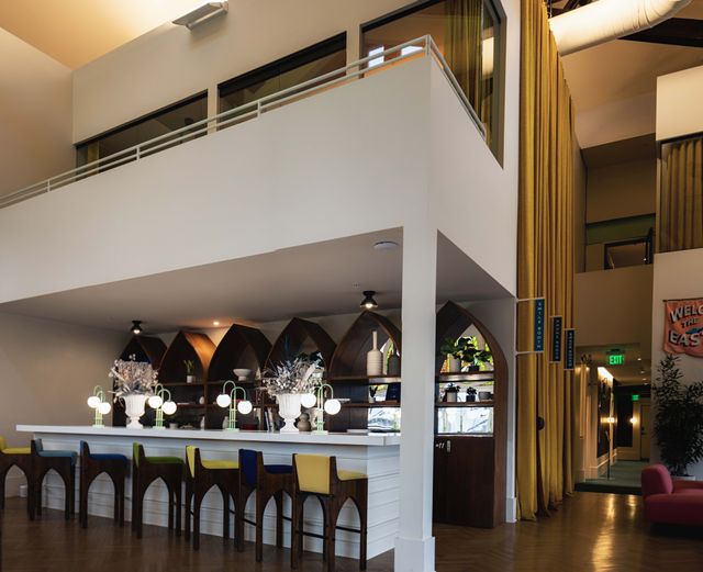 Bright modern hotel lobby bar interior with high mezzanine, arched wooden shelves, colorful bar stools, glowing globe lamps and tall yellow curtains