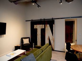Cozy loft-style living room with green velvet sofa, white coffee table, black armchair with patterned pillow, rustic sliding barn door featuring oversized painted letters, exposed industrial ducts and track lighting