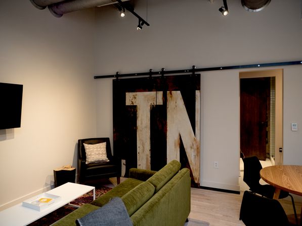 Cozy loft-style living room with green velvet sofa, white coffee table, black armchair with patterned pillow, rustic sliding barn door featuring oversized painted letters, exposed industrial ducts and track lighting