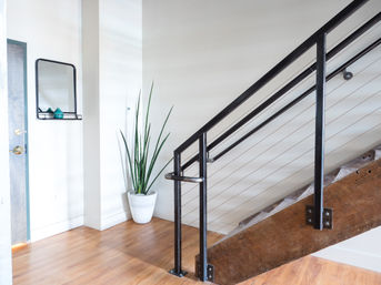 Bright modern entryway with hardwood floors, black metal cable staircase and wooden stringer, tall snake plant in a white pot, and a small wall mirror.