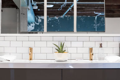 Sleek loft-style bathroom vanity with white subway tile backsplash, two gold faucets, a potted succulent on a glossy white countertop and a rectangular mirror reflecting exposed ceiling beams.