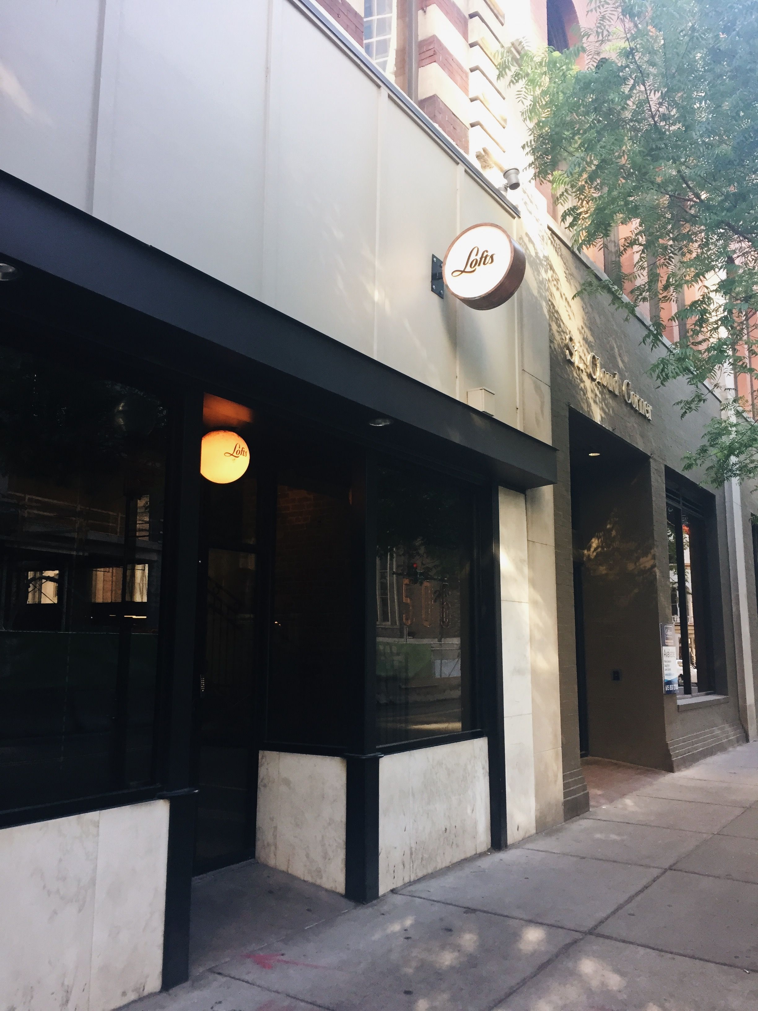 Urban downtown storefront on a tree-lined sidewalk, black-framed glass entrance with marble base, brick upper facade and a round illuminated hanging sign with a script logo, warm evening light.
