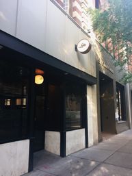 Urban downtown storefront on a tree-lined sidewalk, black-framed glass entrance with marble base, brick upper facade and a round illuminated hanging sign with a script logo, warm evening light.