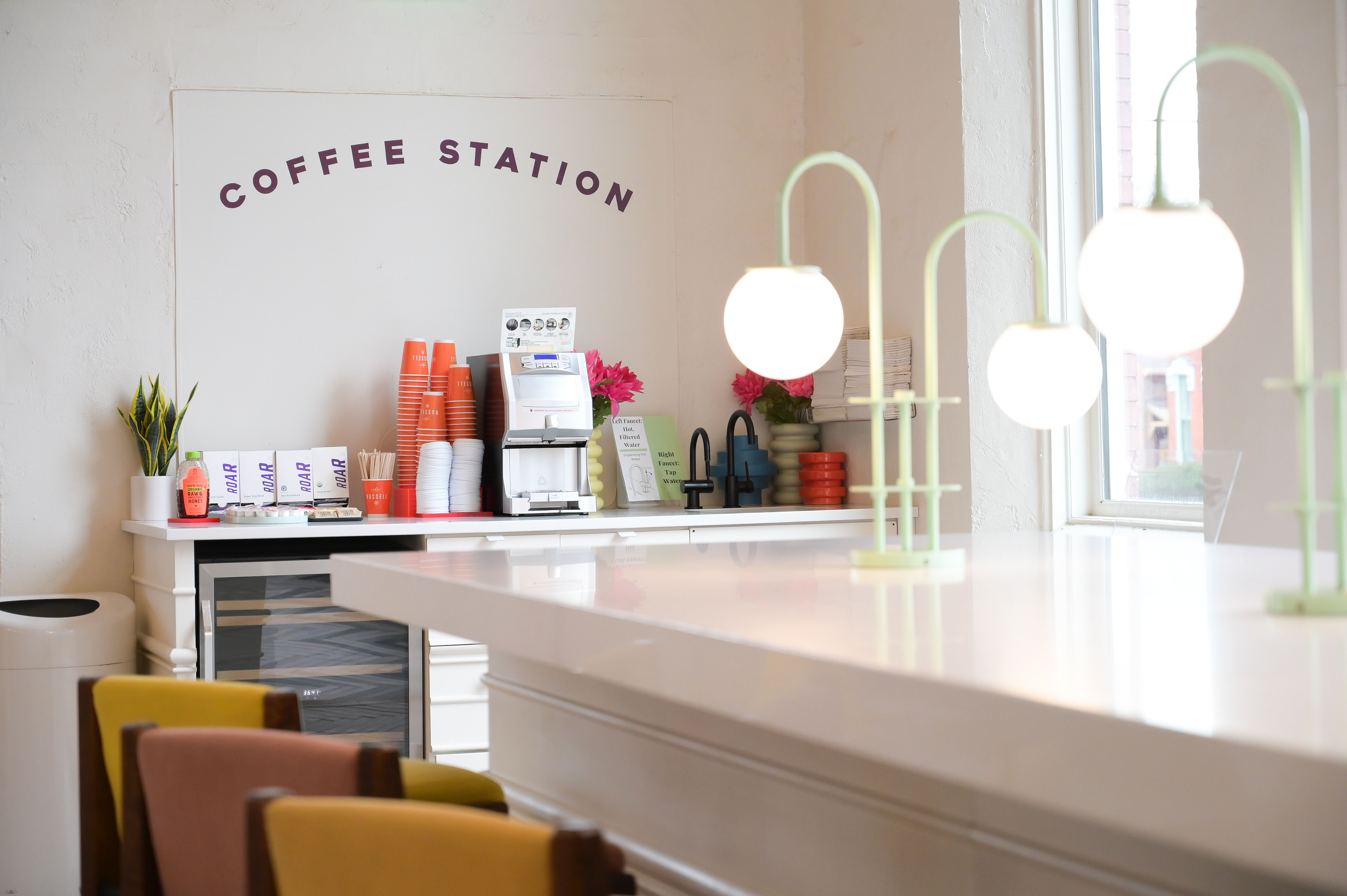 Bright minimalist coffee station in a modern cafe interior with stacked orange takeaway cups, self-serve coffee machine, potted plant and pink flowers, glowing globe pendant lamps, and colorful counter seating by a window.