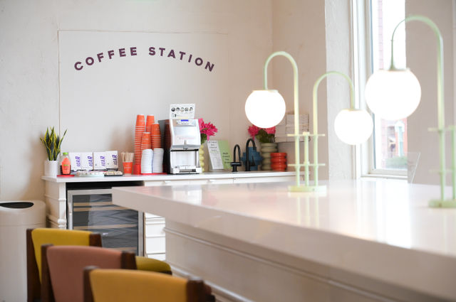 Bright minimalist coffee station in a modern cafe interior with stacked orange takeaway cups, self-serve coffee machine, potted plant and pink flowers, glowing globe pendant lamps, and colorful counter seating by a window.
