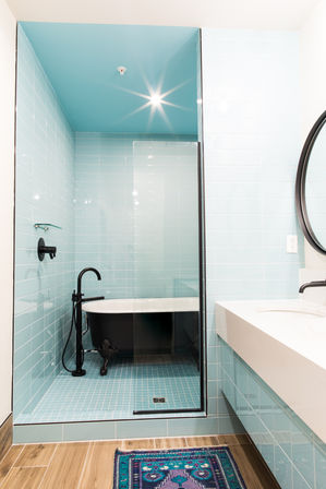 Modern turquoise-tiled bathroom with glass-enclosed shower featuring a black clawfoot tub and matte-black freestanding faucet, wood-look floor and colorful bath rug.