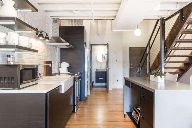 Bright modern urban loft kitchen, open-concept layout with dark wood cabinets, farmhouse sink, stainless steel range and microwave, white subway tile backsplash, breakfast island, hardwood floors, and metal cable-rail staircase.