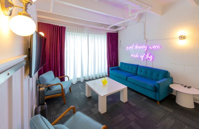 Modern lounge with teal sofa, mid-century blue armchairs, white coffee table, floor-to-ceiling sheers and magenta curtains, neon pink wall sign reading sweet dreams were made of this