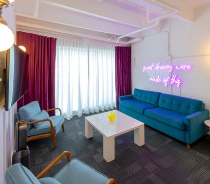 Modern lounge with teal sofa, mid-century blue armchairs, white coffee table, floor-to-ceiling sheers and magenta curtains, neon pink wall sign reading sweet dreams were made of this