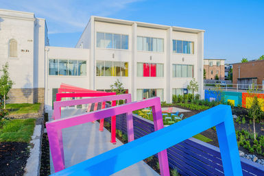 Modern white three-story apartment building overlooking a vibrant urban courtyard with bright pink and blue angled pergolas, colorful outdoor seating, landscaped greenery, and a clear blue sky.