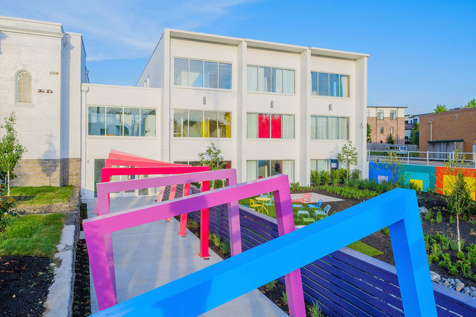 Modern white three-story apartment building overlooking a vibrant urban courtyard with bright pink and blue angled pergolas, colorful outdoor seating, landscaped greenery, and a clear blue sky.