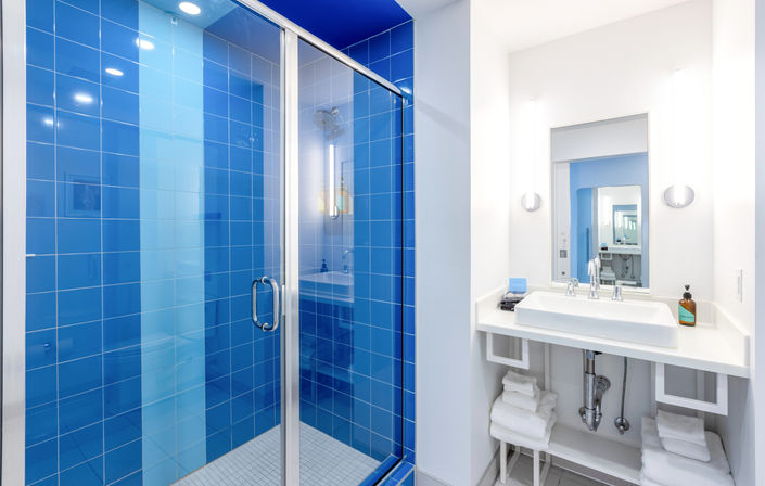 Bright modern bathroom with cobalt-blue tiled walk-in shower behind a glass door, white vanity with rectangular sink, chrome fixtures, wall mirror and neatly folded towels.