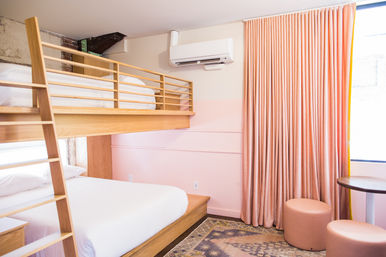 Cozy pastel-pink hotel room with wooden loft bunk bed, crisp white linens, wall-mounted air conditioner, floor-length pink curtains, round table and pouf seating