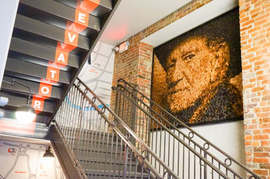 Industrial stairwell with metal railings, black steps labeled ELEVATOR, exposed brick wall and a large pixelated mosaic portrait above a map mural referencing East Nashville, lit by globe pendant lights.