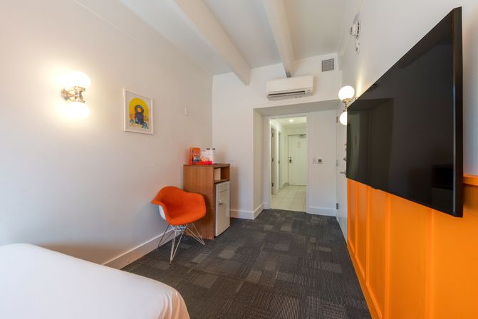 Bright modern hotel room with orange accent chair and panel, wall-mounted flat-screen TV, mini-fridge and wood cabinet, wall sconces and air conditioner above the doorway.