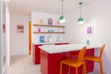 Bright modern office kitchenette with glossy red cabinets, white quartz breakfast bar, two orange bar stools, wood floating shelves, green pendant lights and a coffee station