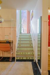 Mint-colored staircase with gold-lettered risers reading 'RISE & SHINE' and 'TODAY IS THE DAY' in a colorful urban boutique-style studio — pink walls, coral desk chair, full-length mirror, and exposed brick visible at top of stairs.
