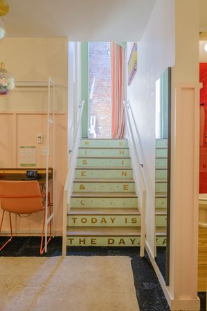 Mint-colored staircase with gold-lettered risers reading 'RISE & SHINE' and 'TODAY IS THE DAY' in a colorful urban boutique-style studio — pink walls, coral desk chair, full-length mirror, and exposed brick visible at top of stairs.