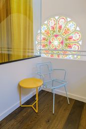 Interior seating nook with a yellow round side table and pastel blue wire chair on dark wood floor, mustard curtain to the left and a colorful stained-glass rose window in the background.