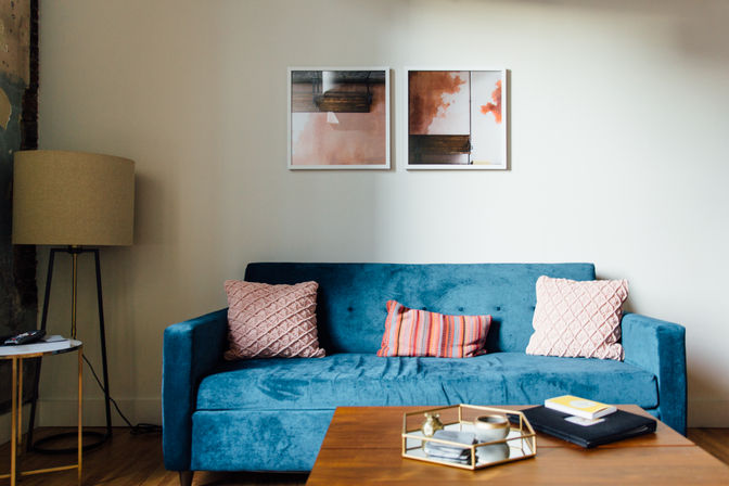 Modern living room with teal velvet sofa, pink textured throw pillows and striped lumbar cushion, wooden coffee table with decorative tray and books, floor lamp and framed abstract wall art.