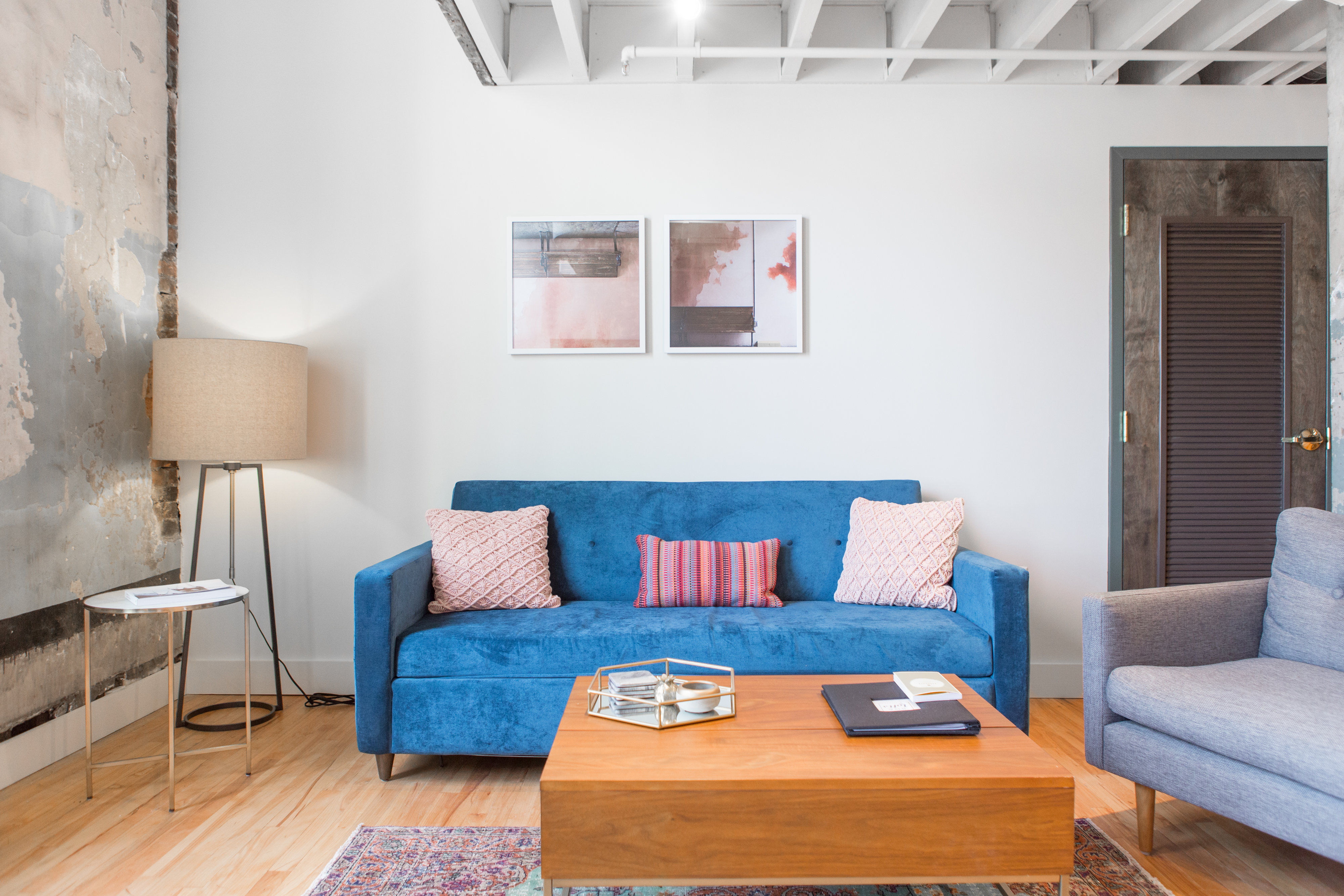 Bright modern loft living room interior with a blue velvet sofa and pink textured pillows, wooden coffee table with decorative tray, tripod floor lamp by a distressed plaster wall, abstract framed art and exposed ceiling beams.