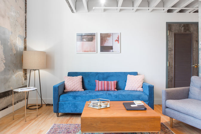 Bright modern loft living room interior with a blue velvet sofa and pink textured pillows, wooden coffee table with decorative tray, tripod floor lamp by a distressed plaster wall, abstract framed art and exposed ceiling beams.