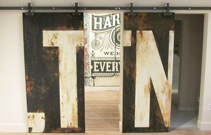 Rustic sliding barn-style doors with oversized distressed painted letters T and N on industrial track, vintage graphic wall visible through center and light wood floor